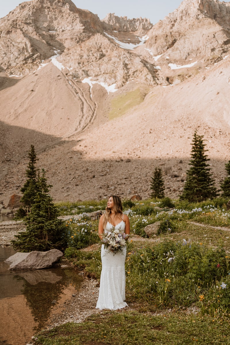 Bride stands next to a calm lake in a wedding dress holding a bouquet. She looks over her shoulder, and the sunlit Colorado mountains are shining behind her.