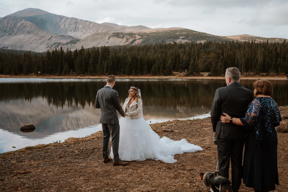 Couple stands on the shores of Brainard Lake at sunset in October for their romantic elopement. The mountains are reflected in the lake behind the couple and two elopement guests stand off to the side.