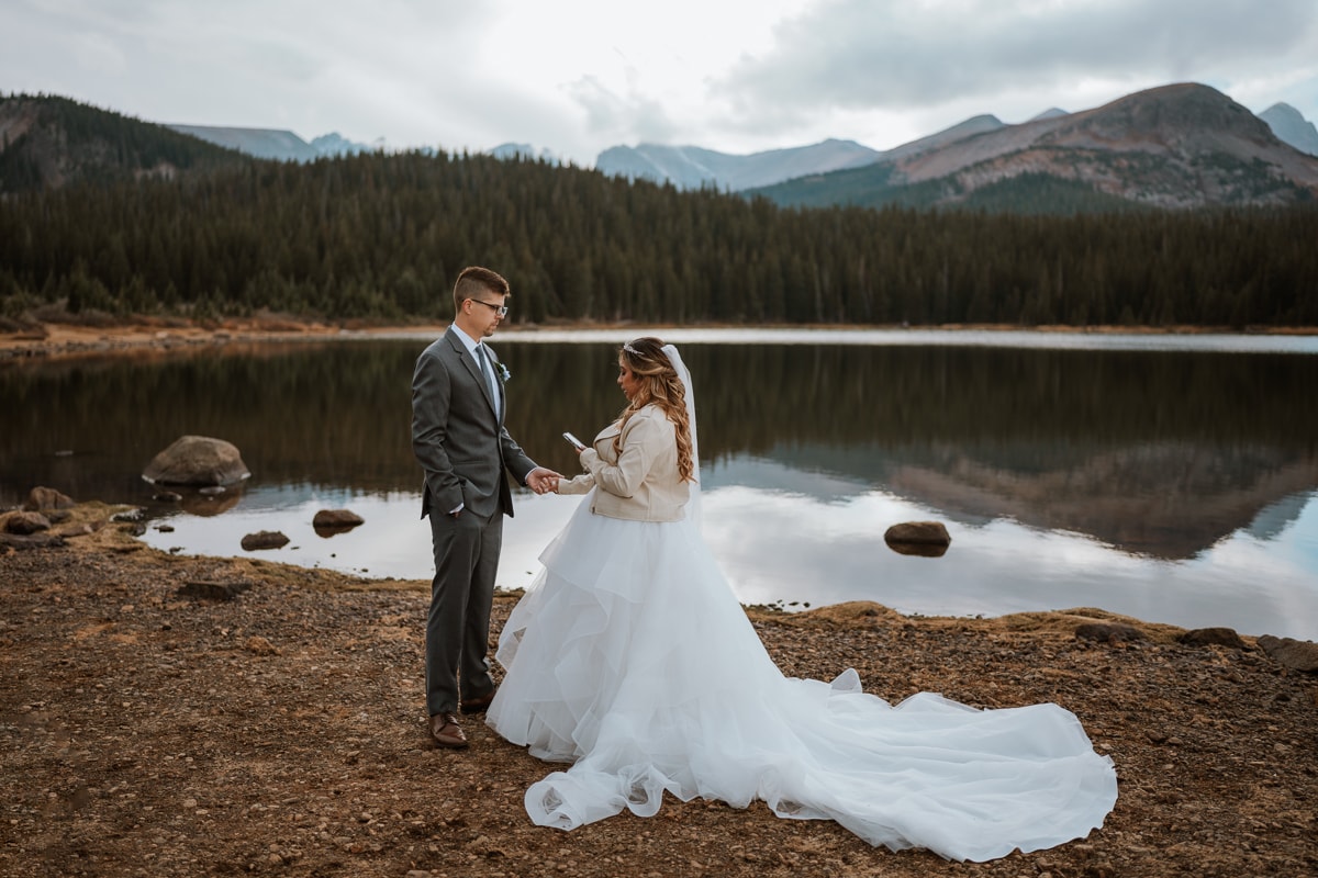 Couple reads their wedding vows at sunset in the Colorado mountains. Brainard Lake is behind the couple and the mountains are perfectly reflected in the lake.