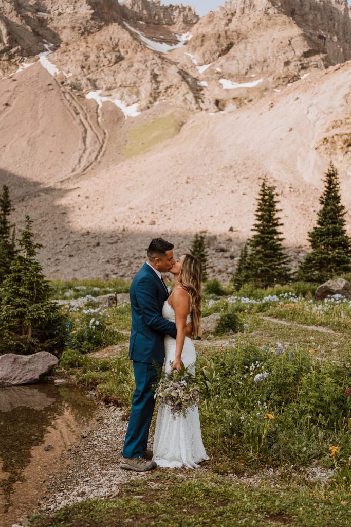 A bride and groom share a kiss on a rock after hiking 4 miles to have their elopement next to a crystal clear blue lake in Colorado.