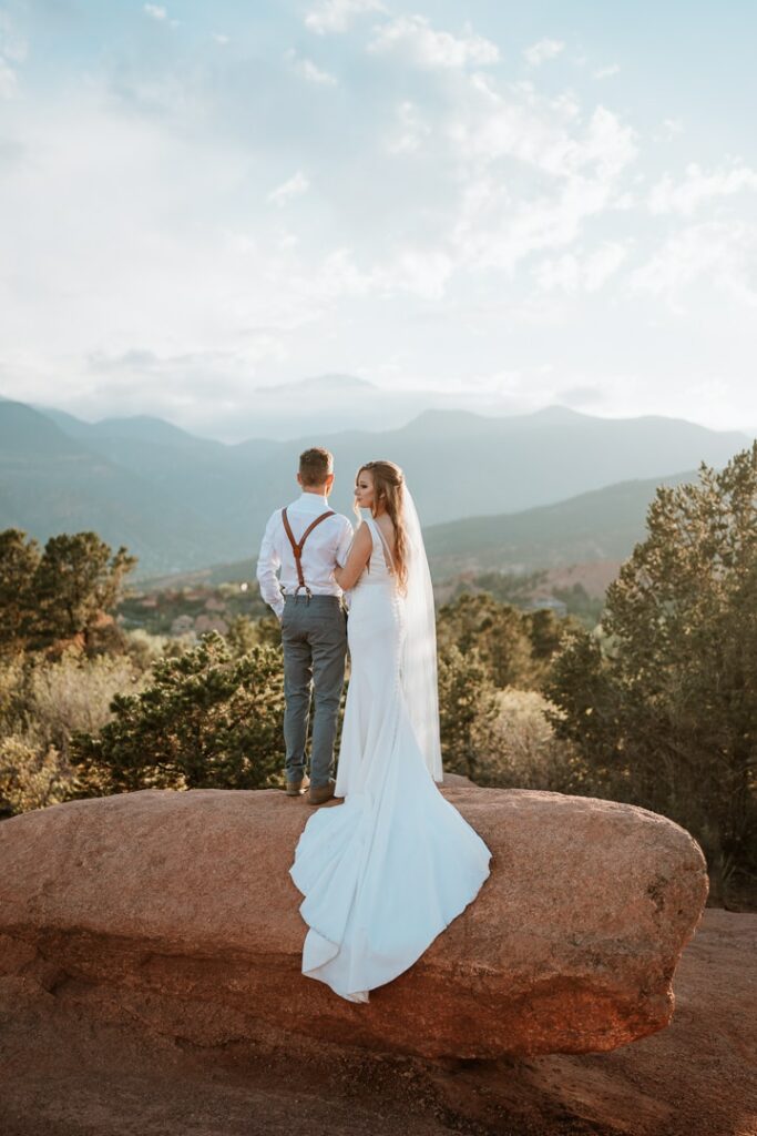 Bride and groom stand on a large red rock formation with Pikes Peak standing tall behind them for their elopement photos in Garden of the Gods.