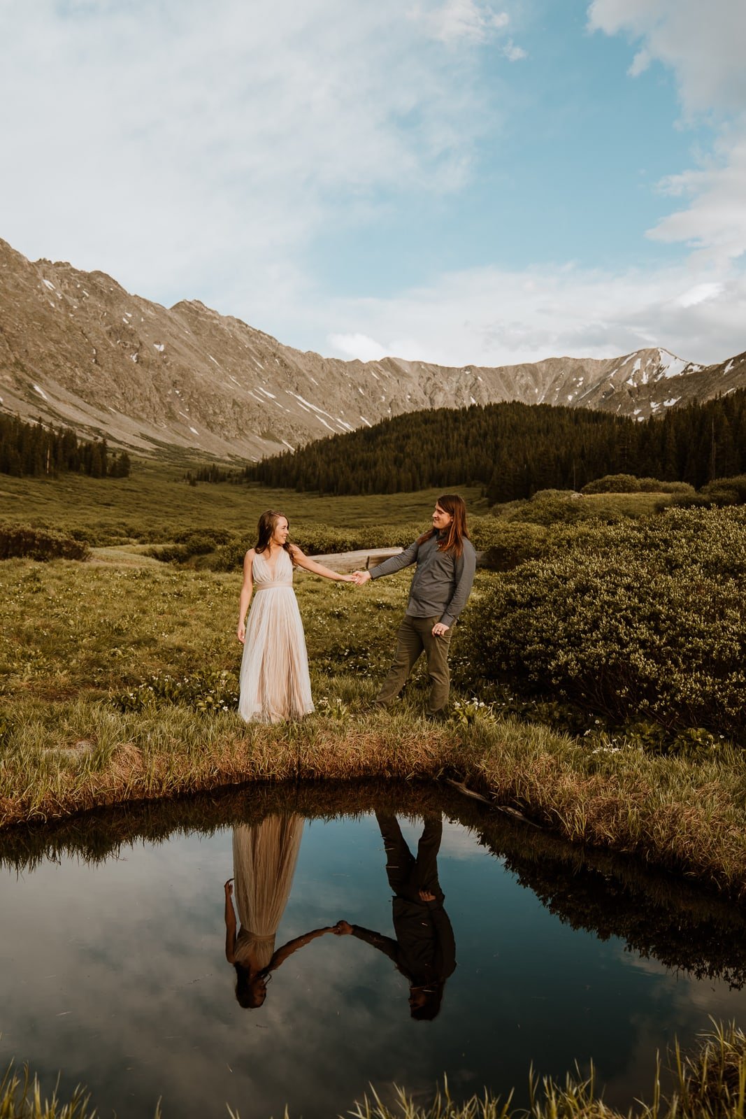 Bride and groom hold hands and walk in front of a calm and reflective lake for their adventurous mountain elopement