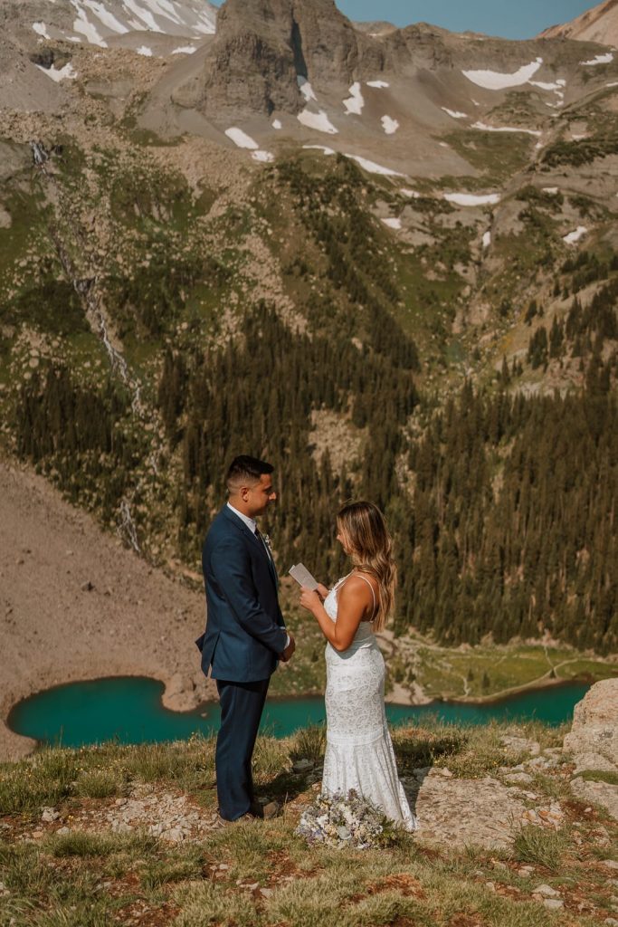 Colorado couple reads their custom elopement ceremony script on a mountain in Telluride with a blue lake behind them.