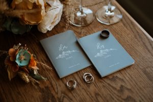 Blue his and hers elopement vow books lay on a wooden table with the couple's wedding rings and bridal bouquet.