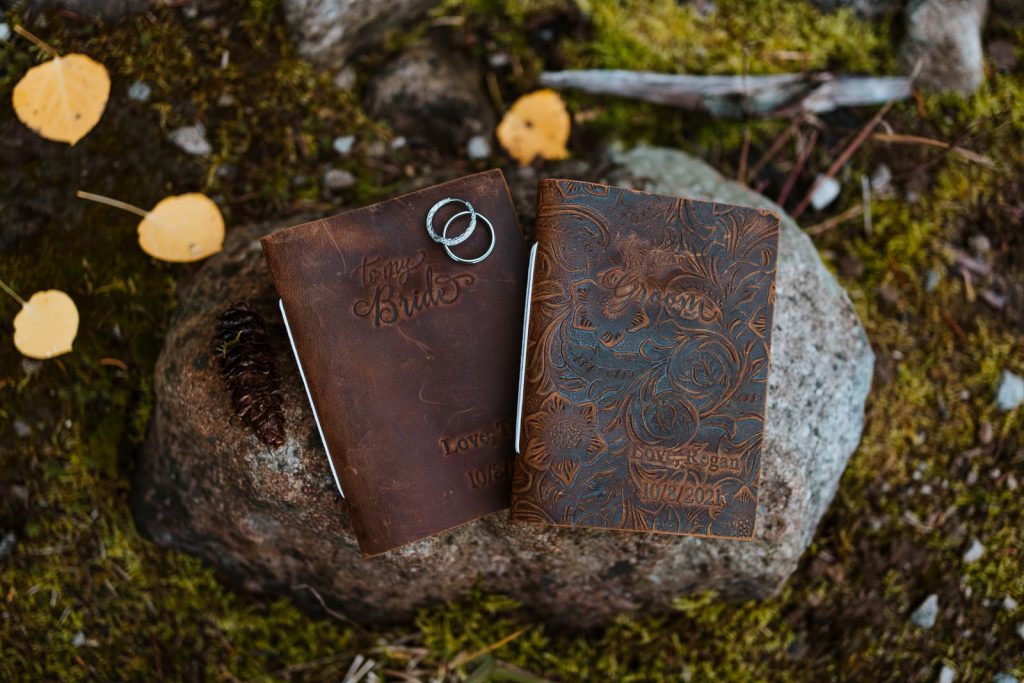 Two leather vow books sitting on a rock next to a pinecone surrounded by aspens.