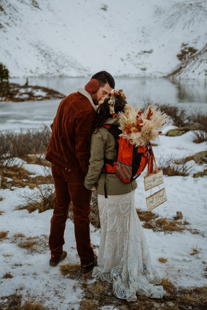 Bride and groom standing in a patch of snow with signs on their backpacks that say 'Just Married'