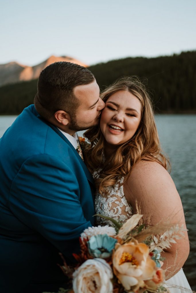 Groom kisses his new wife's cheek as she laughs after they read their elopement vows at a beautiful lake in Colorado that shines behind them in the photo.