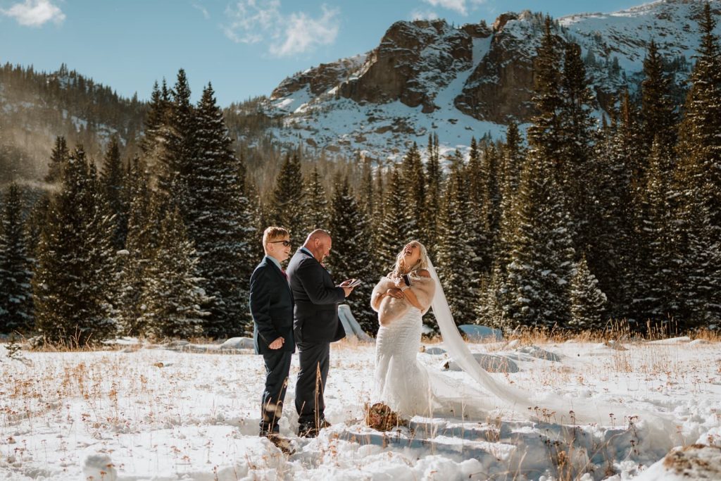 Bride laughs as the groom reads his elopement vows in the snow. The snowy mountains and pine trees are contrasted behind them.