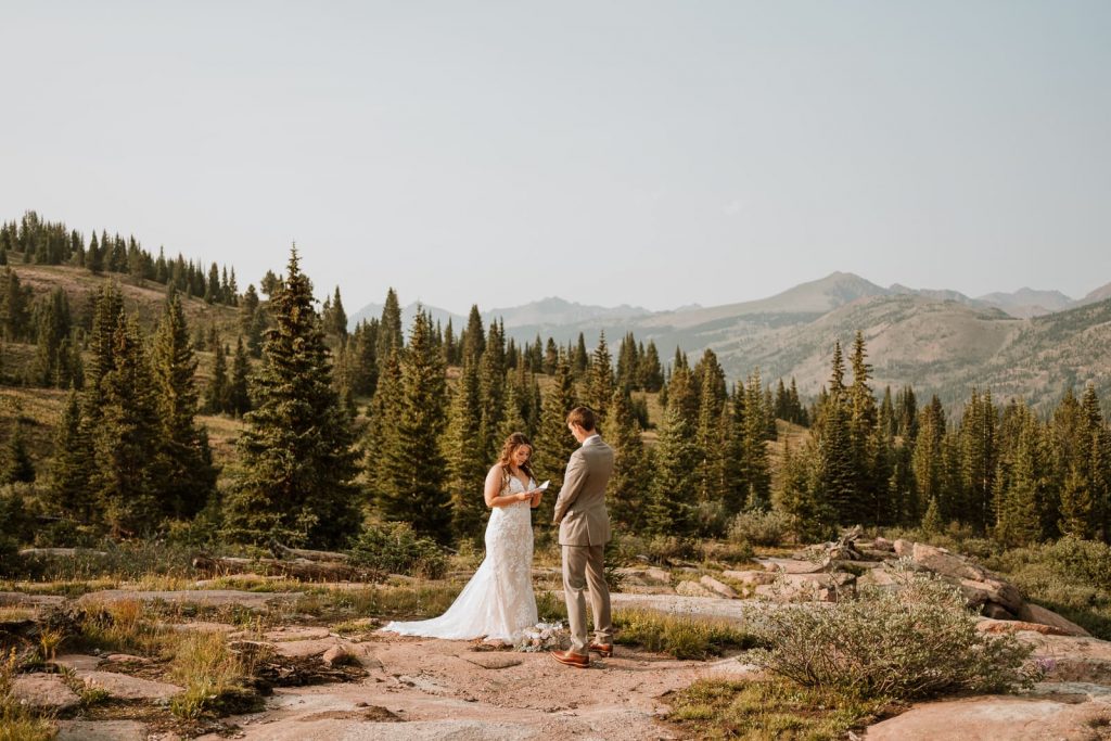 Bride reads her elopement vows to her future husband as they stand on a rock in front of a Colorado mountain with pine trees behind them.