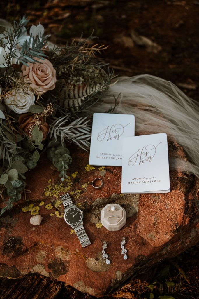A pair of vow books lay on a red rock with the bride's earrings, veil, and bridal bouquet.