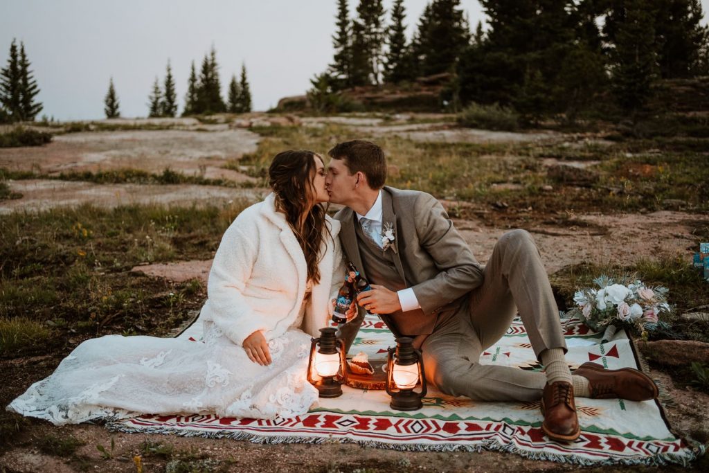 Newly married couple shares a kiss and a beer toast while sitting on a blanket with lanterns after sunset.