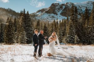 Bride and groom laugh as they read their vows in the snow on a sunny afternoon in Colorado. You can see the wind blowing snow off the pine trees and mountains in the background. Their only guest, their son, stands beside them.