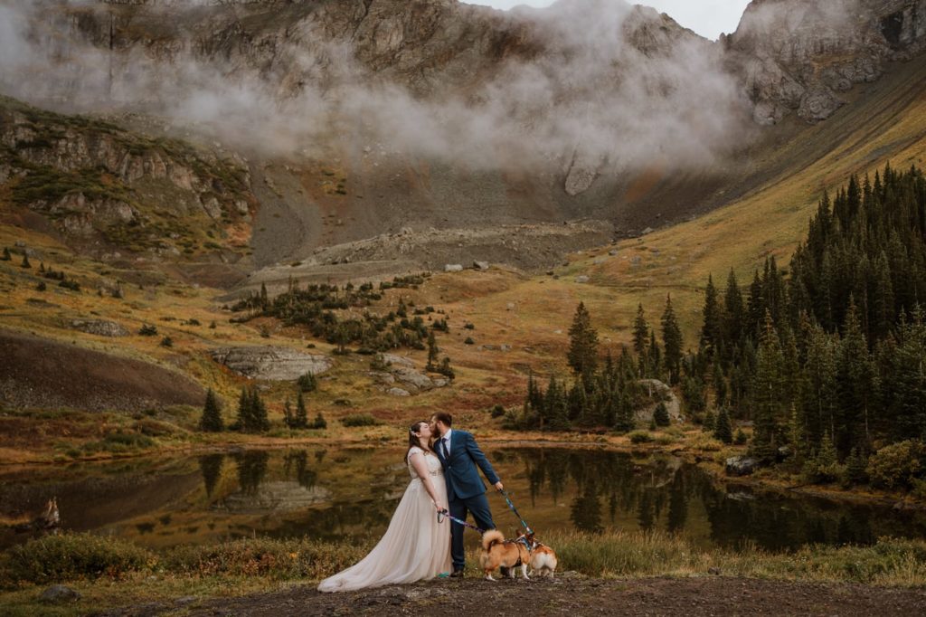 Image of bride and groom kissing while holding their corgi's leashes. They are standing in front of a reflective lake in Telluride, and the mountains and clouds behind the couple are reflected in the lake. Colorado elopement photographer, Paige Weber Photography, captured this image in 2021.