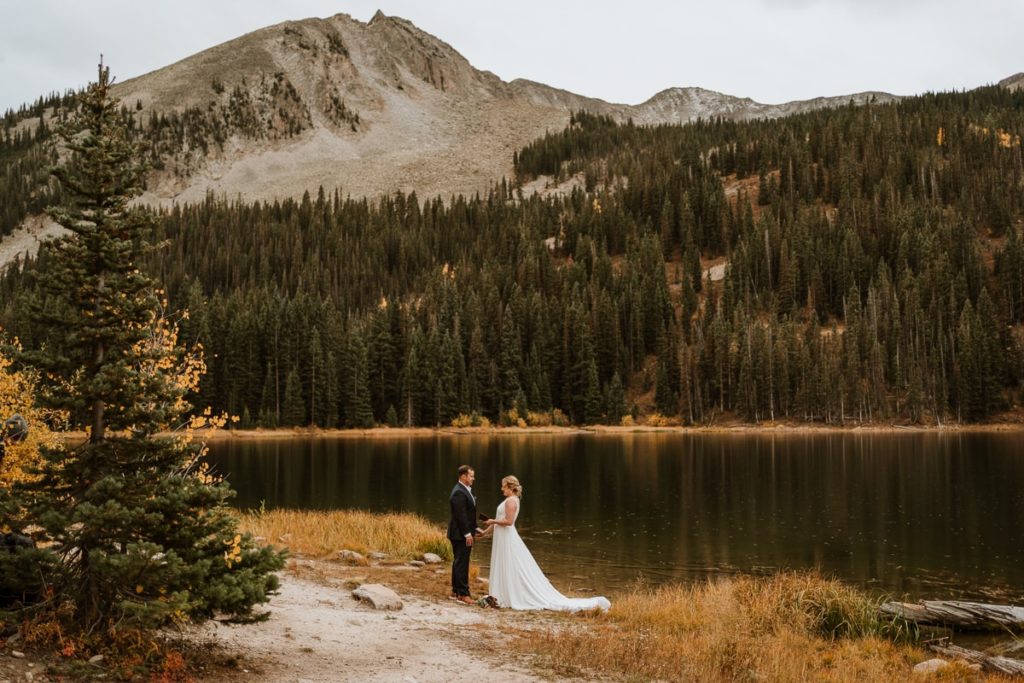 Bride and groom read their wedding vows next to an alpine lake with mountains in the background.