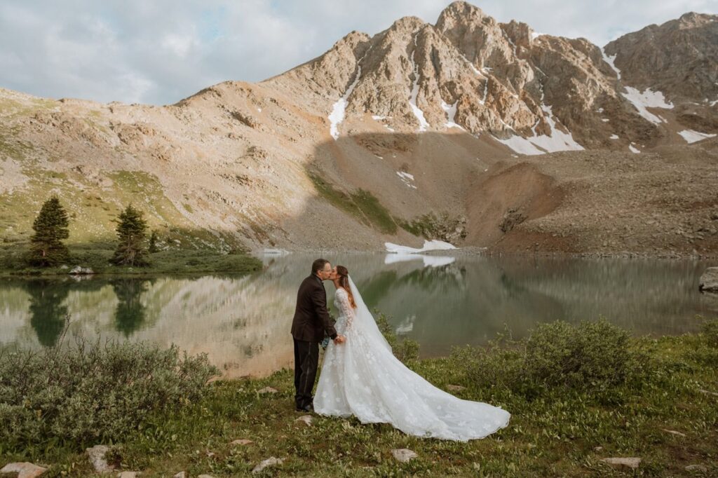 Bride and groom share a first kiss during their mountain elopement after reading their vows in front of a blue reflective alpine lake. A dramatic mountain stands tall behind the couple.