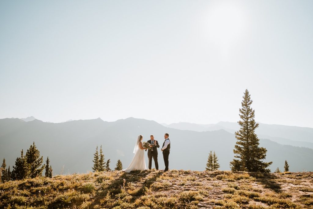 Bride, groom, and their officiant stand on a hill in the late afternoon for a top of the mountain elopement ceremony. It was a hazy day in Colorado due to the wildfire smoke, so the mountains appear as a silhouette behind the couple.