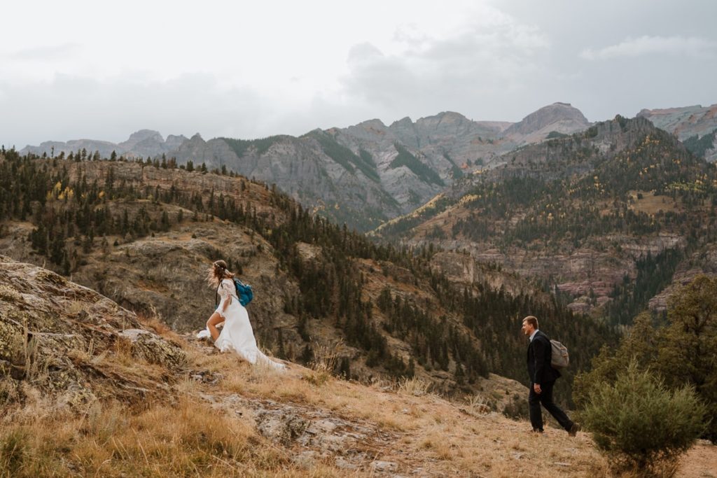 Bride lifts up the front of her dress as she hikes up a steep hill with her husband walking behind her. The mountains of Ouray, Colorado are towering behind them.