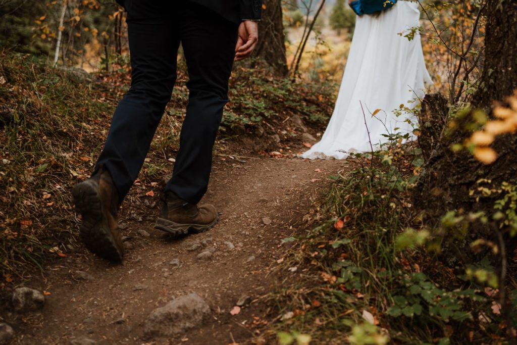 Closeup image of a bride's wedding dress dragging along a hiking trail. Her husband is walking behind her in a suit and brown hiking boots. The trail is surrounded by colorful foliage as this mountain elopement took place in Ouray Colorado.