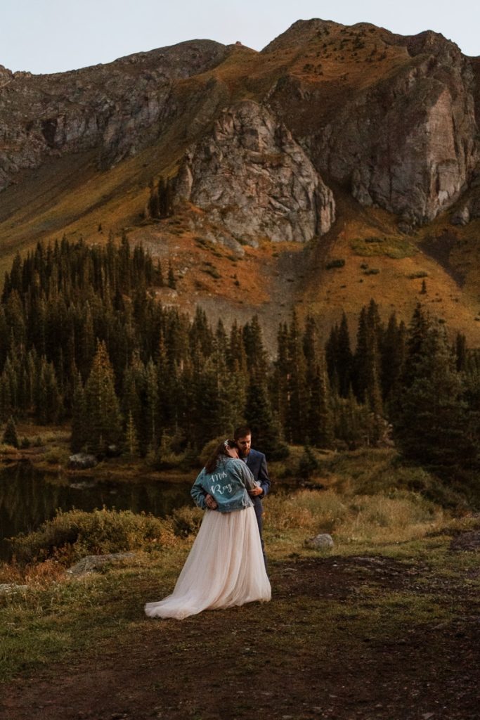 Bride and groom share their first dance during blue hour. She is wearing a jean jacket over her wedding dress and laying on his shoulder as the mountains and pine trees tower behind them.