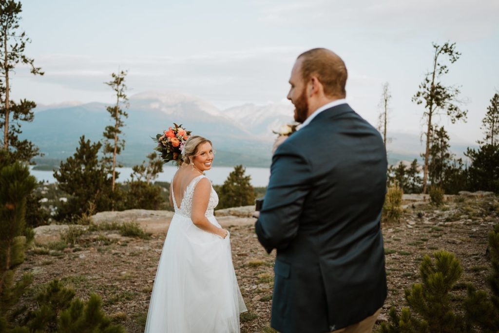 Bride smiles as she looks back at her groom. They are walking to their sunrise picnic in the distance for their mountain elopement. The 12,000 foot mountains and a lake are off in the distance behind the bride.