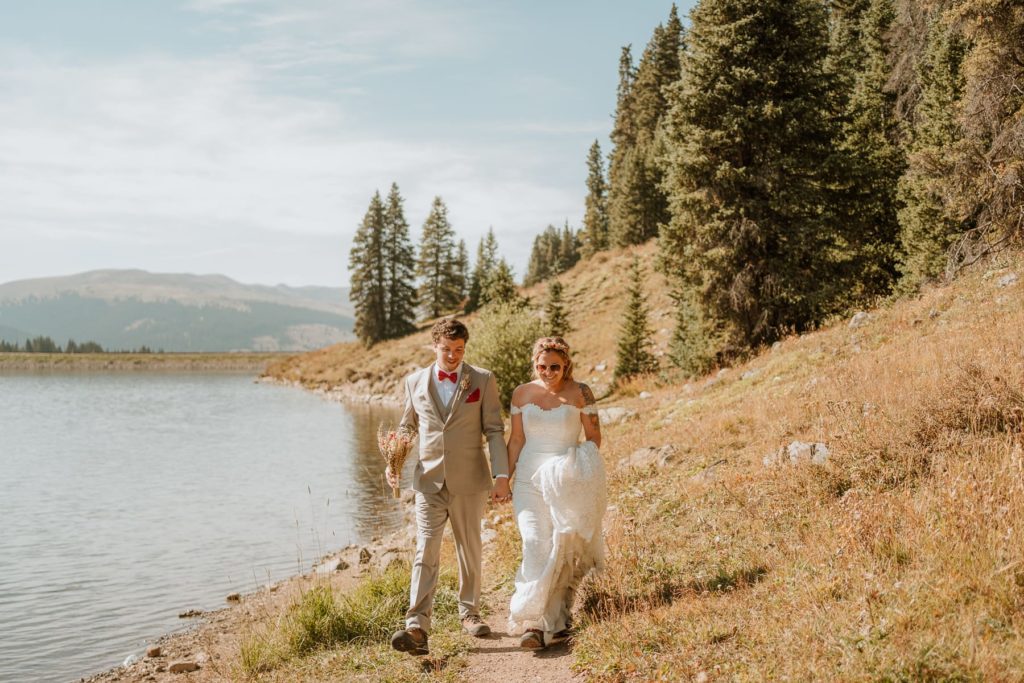 Bride and groom walking hand in hand in their wedding attire on a hiking trail. The sun is bright as it's mid-day, and the bride is wearing heart shaped sunglasses and wedding hiking boots with her wedding dress.