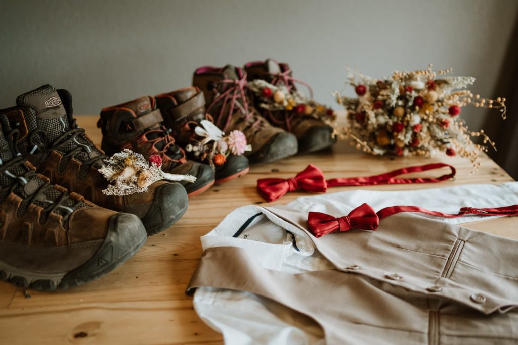 A bride and groom lined up their wedding hiking boots in a row with their son's tiny hiking boots. In the frame to the right is a dried wildflower bouquet, and two matching red bow ties lay in front of the boots with wedding attire.