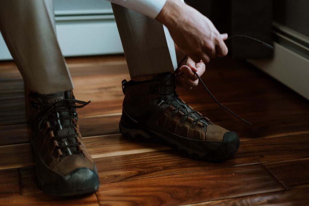 Groom laces up his brown wedding hiking boots. He's wearing a tan suite and the side lighting from the window on the right gives the boots an artistic glow.