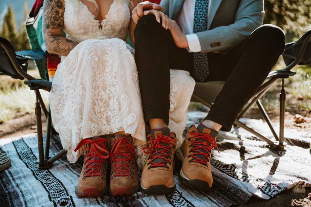 Bride and groom sitting in a camping chair holding hands. You can't see their faces but you can see their wedding attire and matching brown wedding hiking boots with red laces. Photo by Paige Weber Photography, a Colorado elopement photographer.
