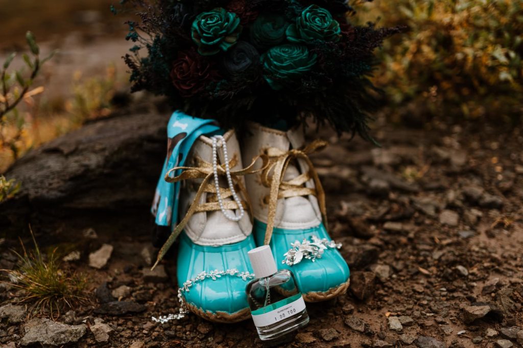 A bride's funky sperry boots sit next to a log with her jewelry, perfume, blue sola wooden bridal bouquet, and her custom hiking socks that have her dog's face on them.