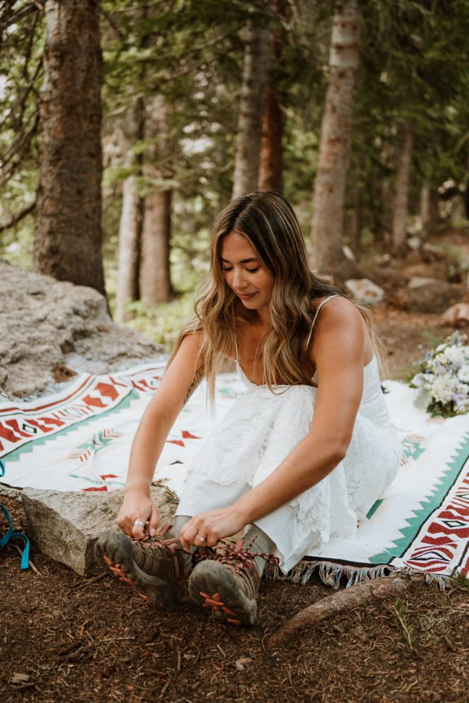 Bride sits in her white wedding dress on a decorated blanket on the ground. She is in the trees and putting on her wedding hiking boots before her first look with her future husband. Photo taken by Paige Weber Photography, a Colorado elopement photographer.