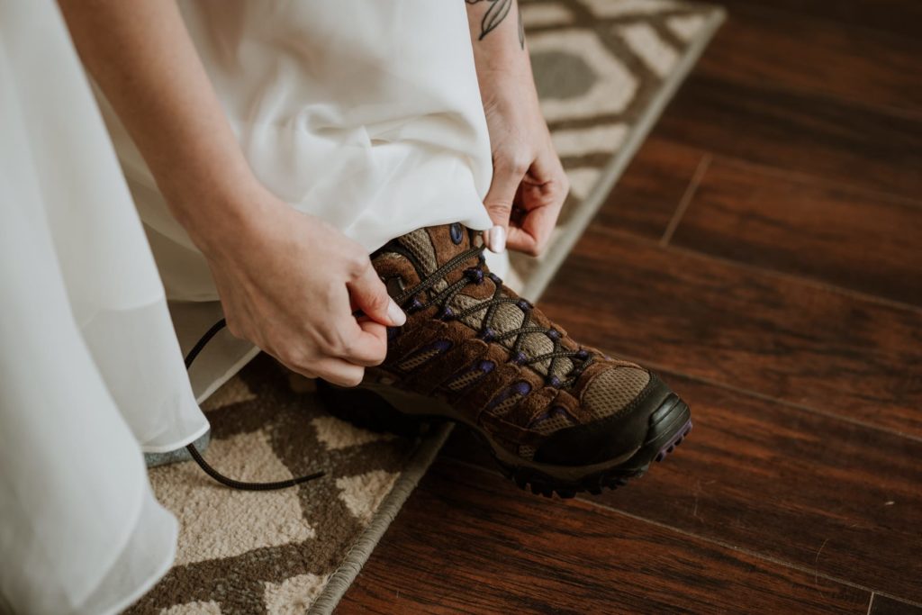 Bride laces up her brown wedding hiking boots. Her wedding dress is laying over the boots to show that she was wearing a long dress.