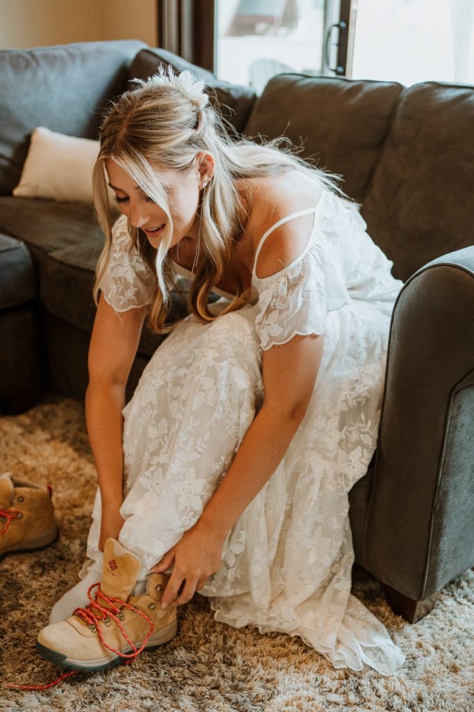 Bride sitting in the living room in her wedding dress. She's on the couch, leaning over and tying her wedding hiking boots.