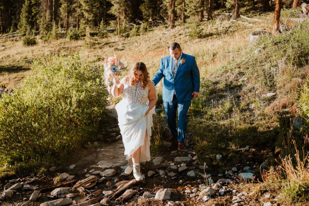 Bride and groom dressed up in wedding attire. They are walking on a hiking trail with trees behind them. The bride is holding up the front of her dress to show off her wedding hiking boots as she crosses a river. The groom is right behind her in a blue suit watching. Photo taken by Paige Weber Photography, a colorado elopement photographer.