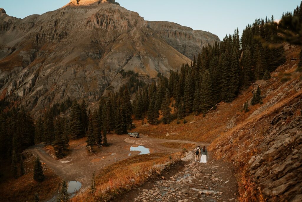 The image shows Katie and Alex, a couple who just had an Ouray elopement, walking down a dirt road on the right side of the frame. The beautiful mountains of Ouray tower behind them with the alpenglow lighting up just the very top of the mountains. The couple's SUV waits at the bottom of the dirt road surrounded by pine trees. The sun is setting and everything in the scene has a warm glow to it.