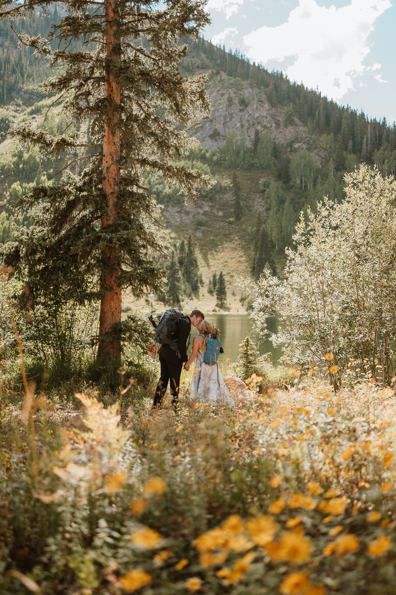 Bride and groom stand next to a lake in Colorado. They are holding hands and leaning in for a kiss. The wildflowers are completely covering the foreground and the mountains tower up behind them. This is the kind of elopement you can expect with your Colorado elopement package.