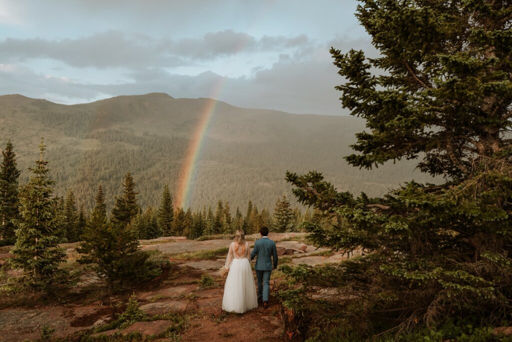 Bride and groom walk on a flat rocky surface towards a vivid rainbow in the distance. The rainbow is overshadowing them and appears right in front of the massive Colorado mountain.