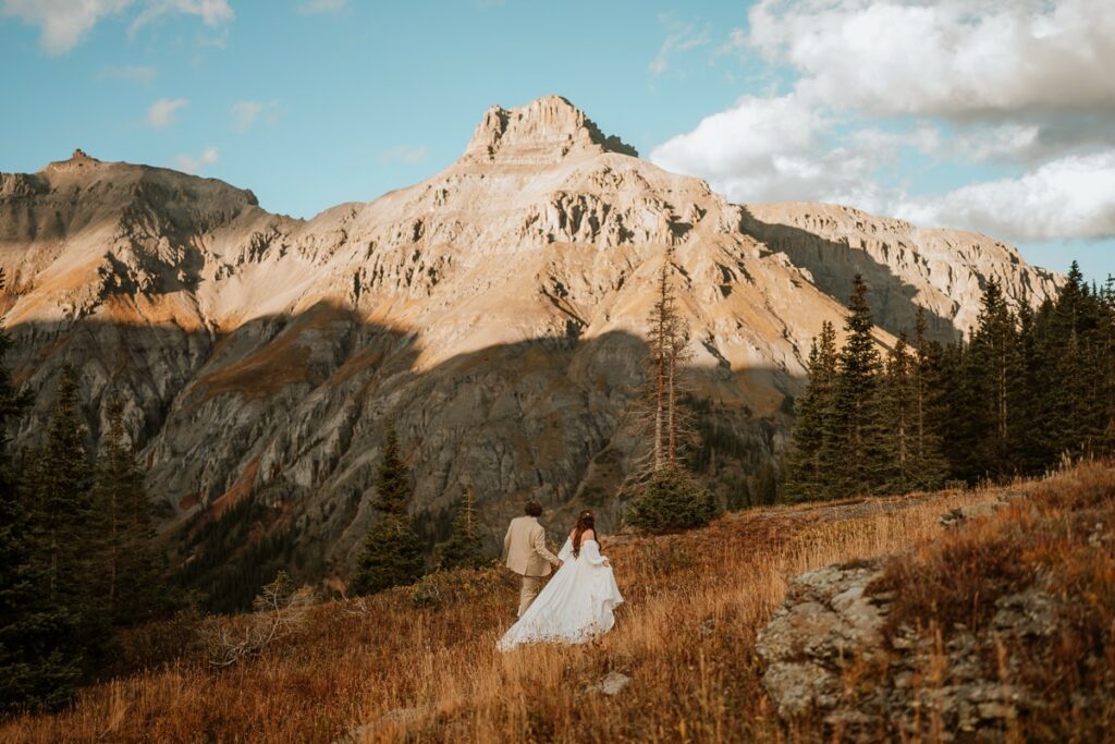 A newlywed couple in wedding attire walk through a field in an alpine basin in Ouray, Colorado. The sunset casts alpenglow on the 13,000 foot mountain in front of them.