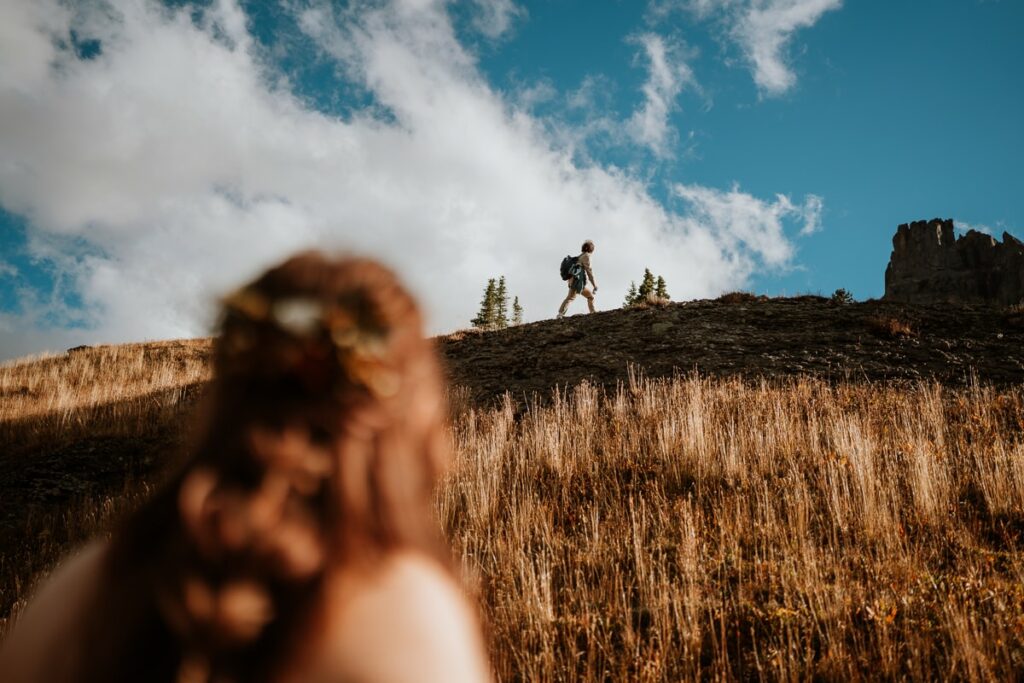 Groom hiking on the mountain-side with his bride watching him in the foreground. White cumulonimbus clouds appear behind him and make his silhouette stand out. Their main goal of their Ouray elopement was to explore, and hiking was the perfect start to that.