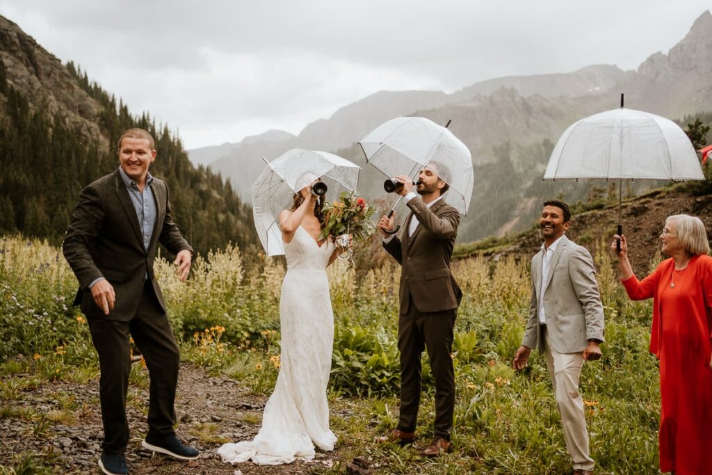 Eloping couple drinks champagne right from the bottle. They are holding clear umbrellas because it just started to rain for their Ouray elopement.