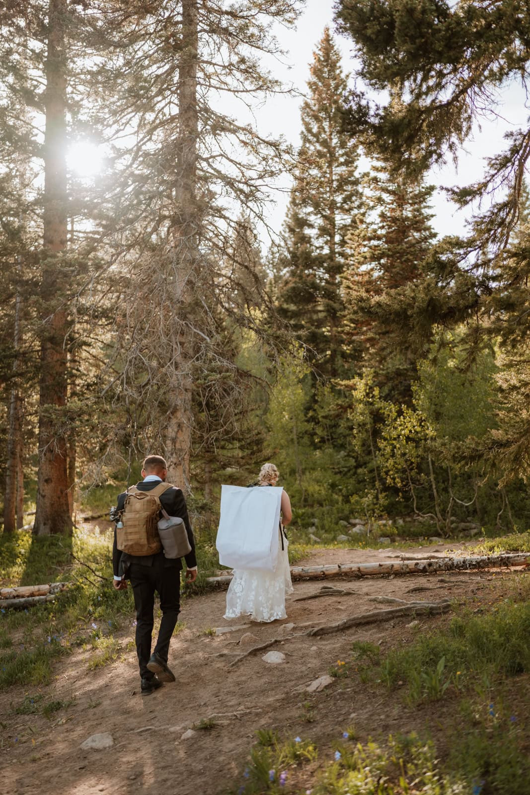 Bride and groom hike on a trail with their backpacks on towards their ceremony location. The trees are tall around them and the sun is shining through the trees.