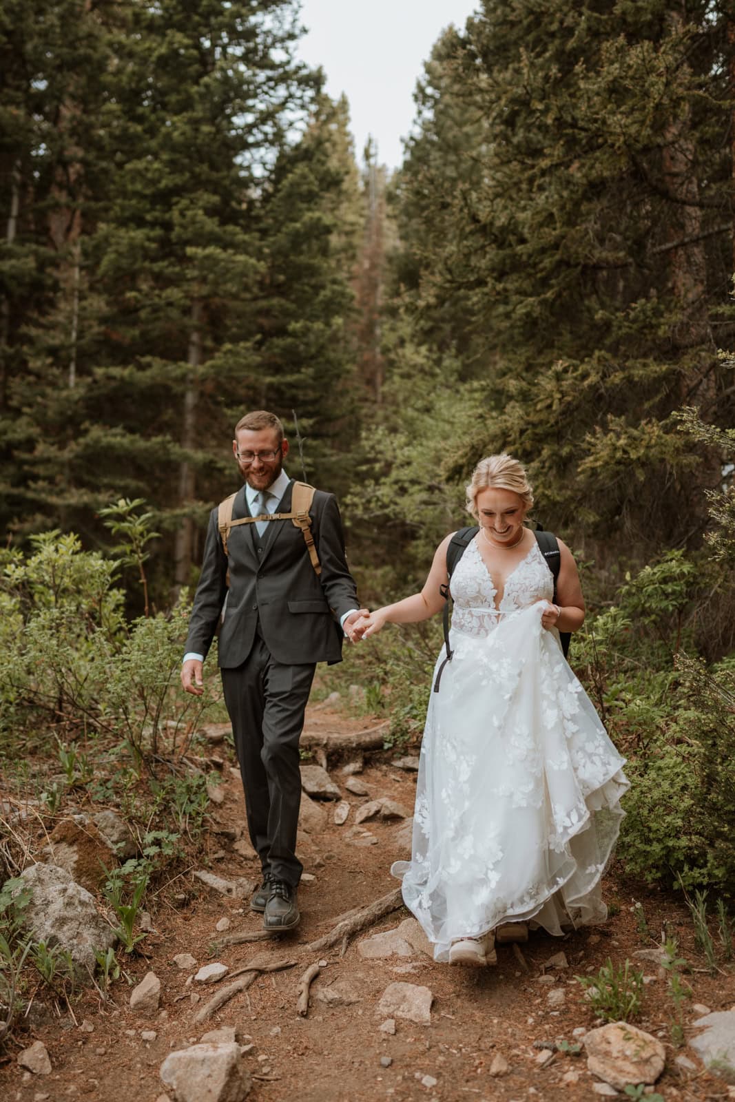 Bride and groom hold hands as they walk down a hiking trail for their hiking elopement in Crested Butte. The bride has a huge smile on her face. She is also holding up her dress so she doesn't trip on it.