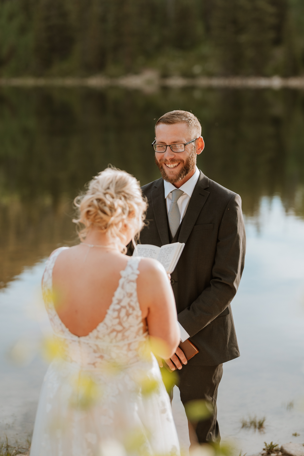 The groom has a huge smile on his face as the bride reads her vows to him. The sunset is casting a beautiful reflection on the lake behind him.