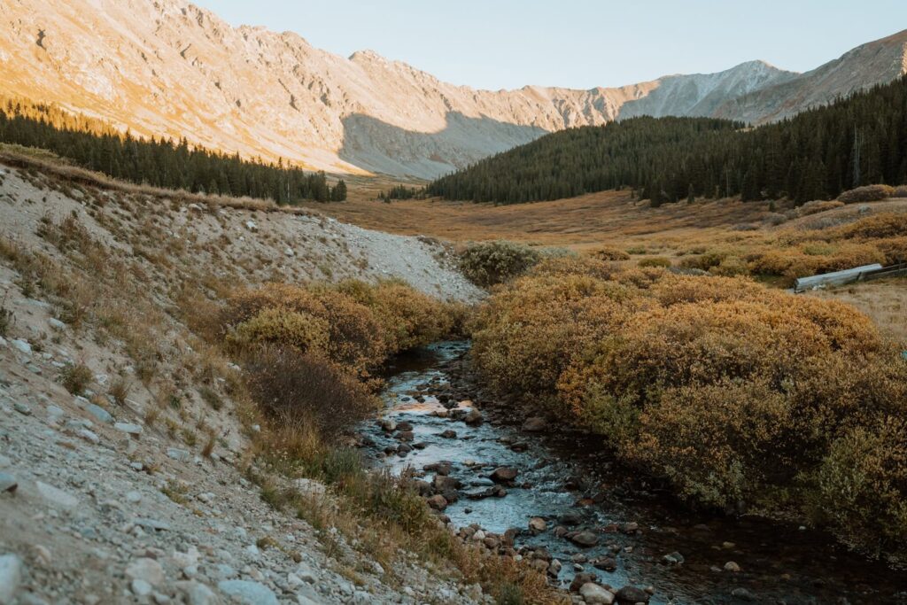 Wide photo that shows a reflective creek that runs through the alpine basin. The alpenglow shines bright on the mountains behind the river.