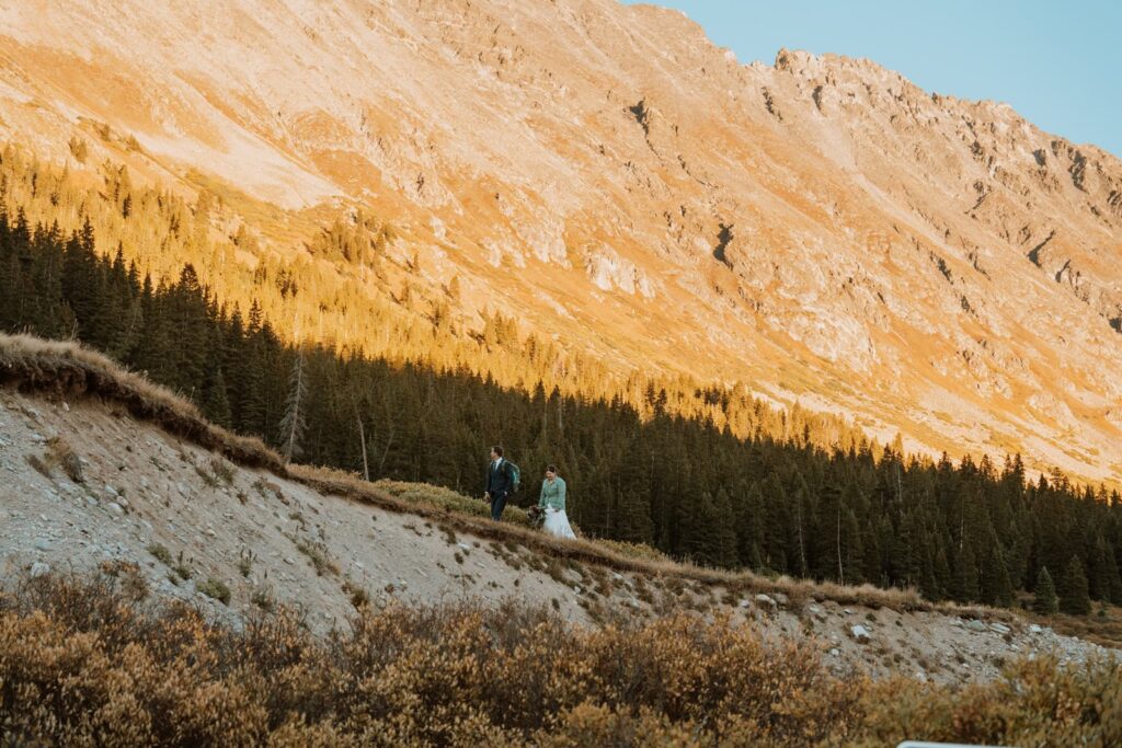 The newlyweds look tiny as they walk along a ridge at sunset. The alpenglow is vivid and orange on the mountains behind them for their small mountain wedding in Colorado.