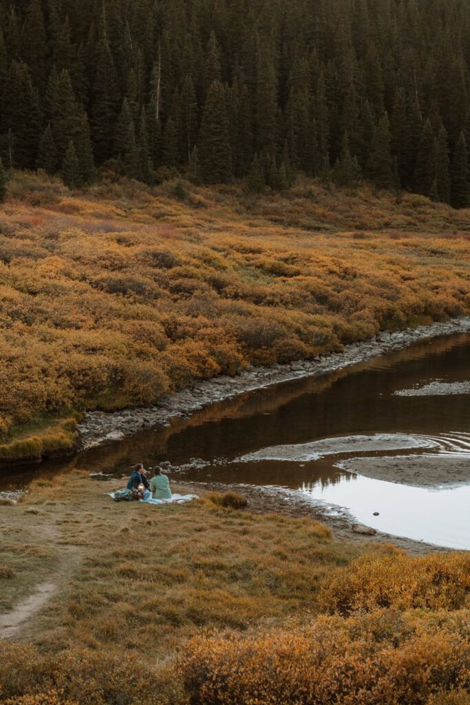 The bride and groom look tiny as they have a sunset picnic next to an alpine lake in Colorado.
