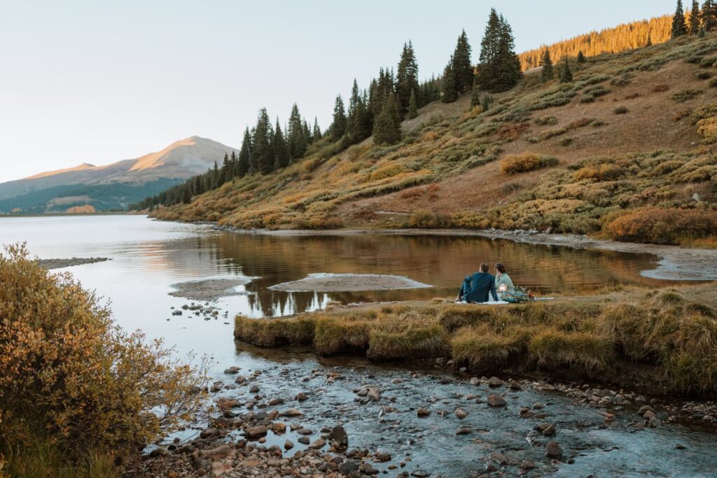 Dramatic photo that shows the vast landscape near an alpine lake by Breckenridge, Colorado. The bride and groom are having a romantic picnic next to the water as the sun sets.