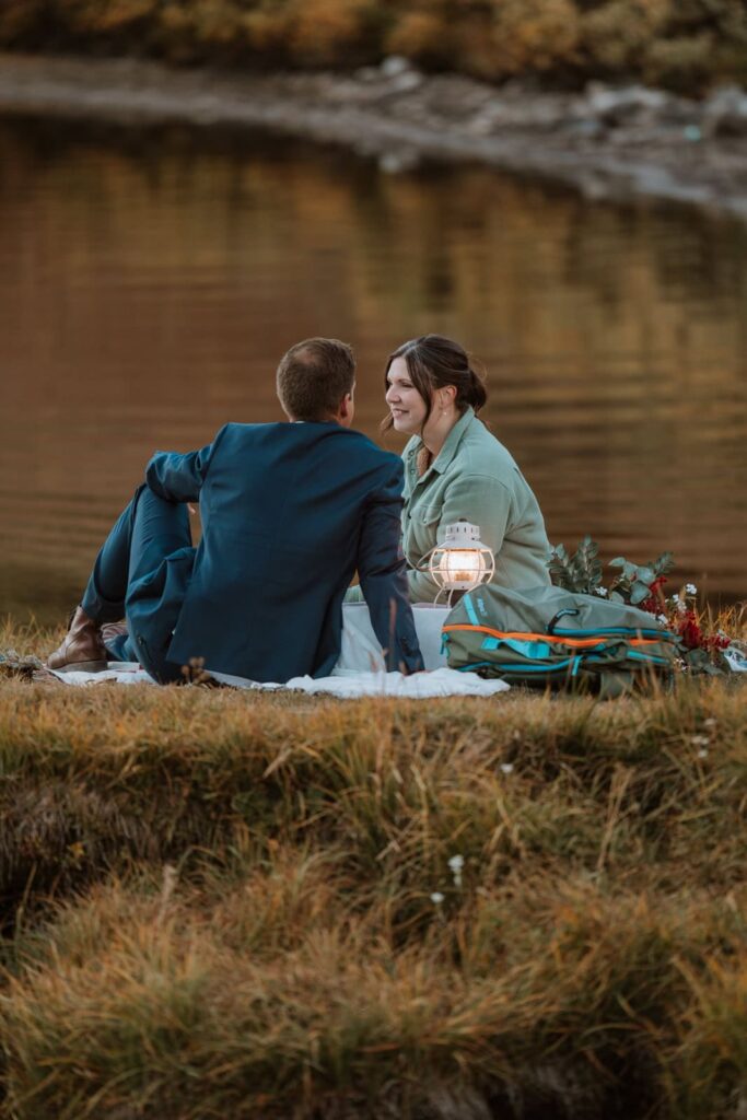 The bride smiles at her groom as they sit next to each other during their romantic sunset picnic. The lake is calm behind them and the white lantern shines brightly in the frame.