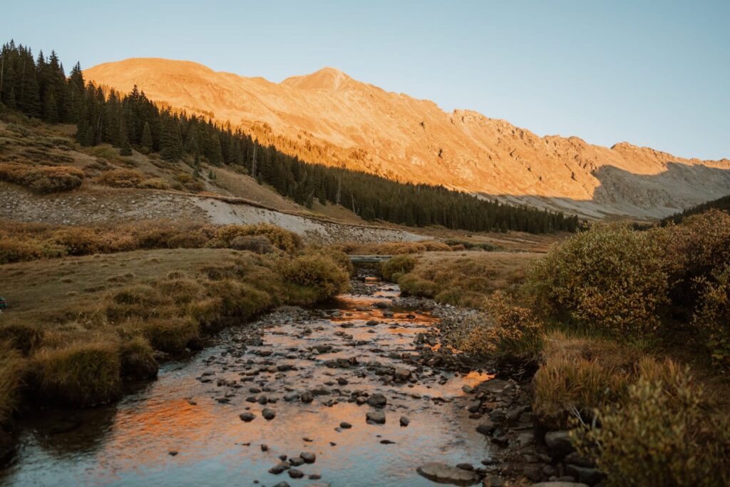 Wide photo that shows sunset at Clinton Gulch reservoir in Colorado. The alpenglow is so bright and vivid that it reflects back in the flowing river in the frame.