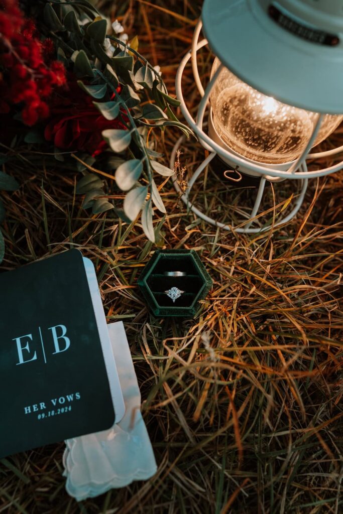 Detail photo of a bride and grooms wedding ring sitting in an Octagon shaped ring box next to a white lantern and dark vow books with the couple's initials.