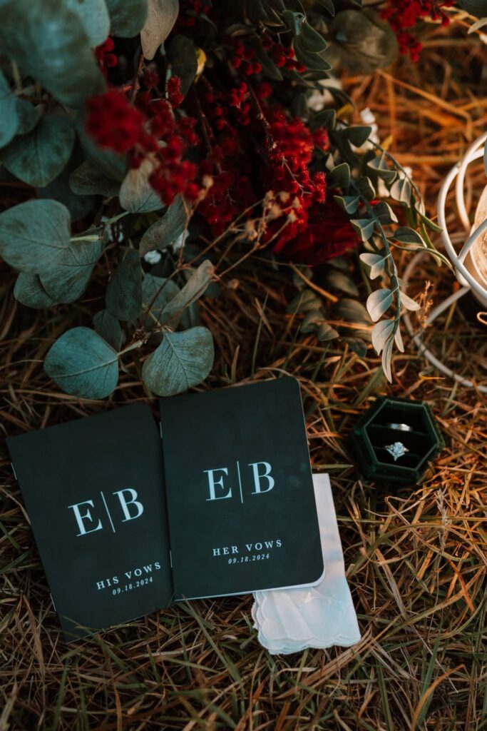 Detail photo shows two dark green vow books with the bride and grooms initials and their wedding date. The vow books sit next to the bride and grooms rings, bouquet and a white lantern.